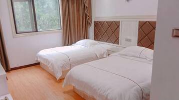 A room with two single beds, a window with curtains, a wooden floor, and a wall-mounted light switch.