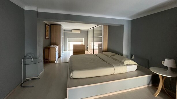 A modern bedroom with a large bed, a bedside table, and a chair. There is a wardrobe and a desk in the background. - [ISOLA] Loft Taramelli (Milano)