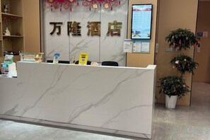 Una recepción de hotel con mostrador de mármol, zona de recepción con tablón de anuncios y plantas en macetas.