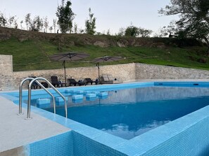Ein Pool mit Liegestühlen und Sonnenschirmen, umgeben von Steinmauern und einem grasbewachsen Hügel.