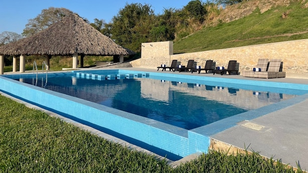 Una piscina rectangular con una estructura de techo de paja, sillas de descanso y un muro de piedra.