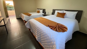 Habitación de hotel con dos camas, cada una con una manta marrón y ropa de cama blanca. Hay una mesita de noche con una lámpara, un equipo de aire acondicionado en la pared y vistas al exterior a través de una ventana.