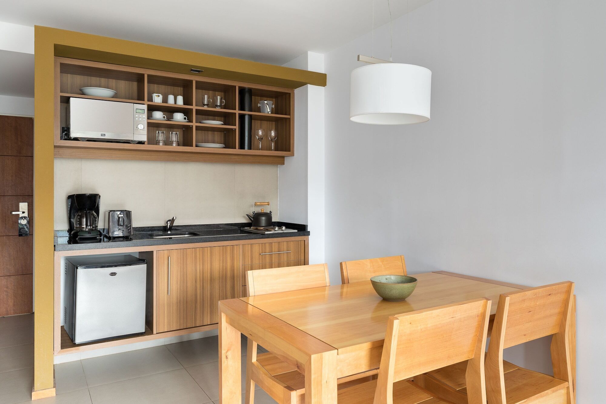 A compact kitchen with a built-in refrigerator, coffee maker, and kettle. There is a wooden dining table with chairs and a bowl on it.