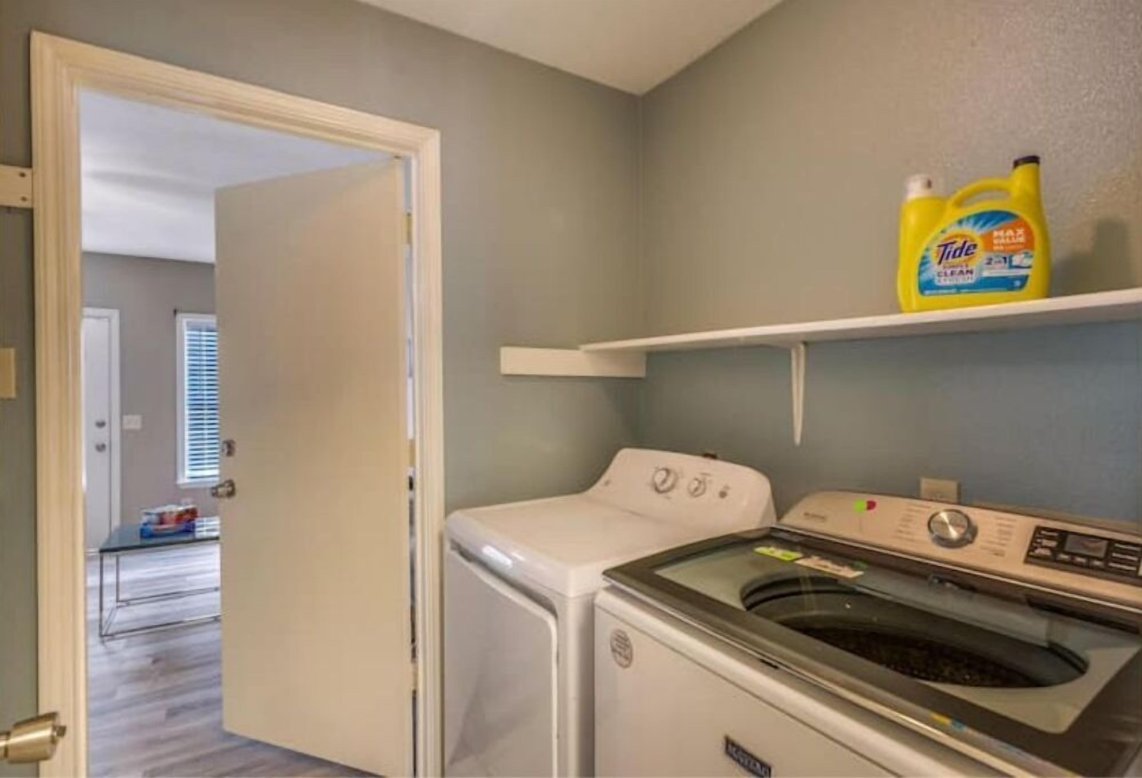 A laundry room with a front-loading washer and dryer, a shelf with Tide detergent, and a door leading to another room.
