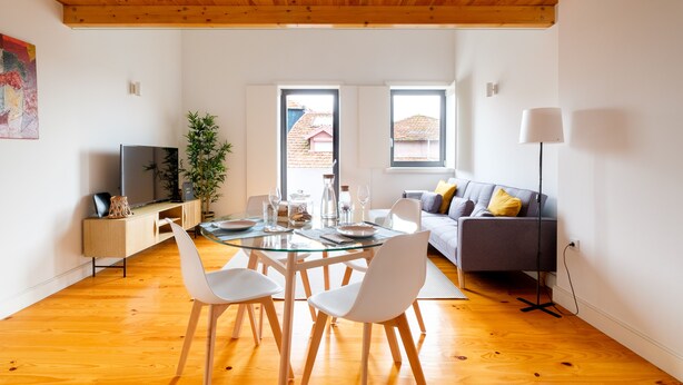 Ein zeitgemäßer Wohnraum mit einem Glastisch, weißen Stühlen, einem grauen Sofa und einem Fernseher auf einer Holzkommode.