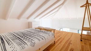 Ein modernes Schlafzimmer mit schrägverdeckter Decke, Holzboden, einem Bett mit gemusterten Bettbezügen, einem Nachttisch und einer Kleiderstange.