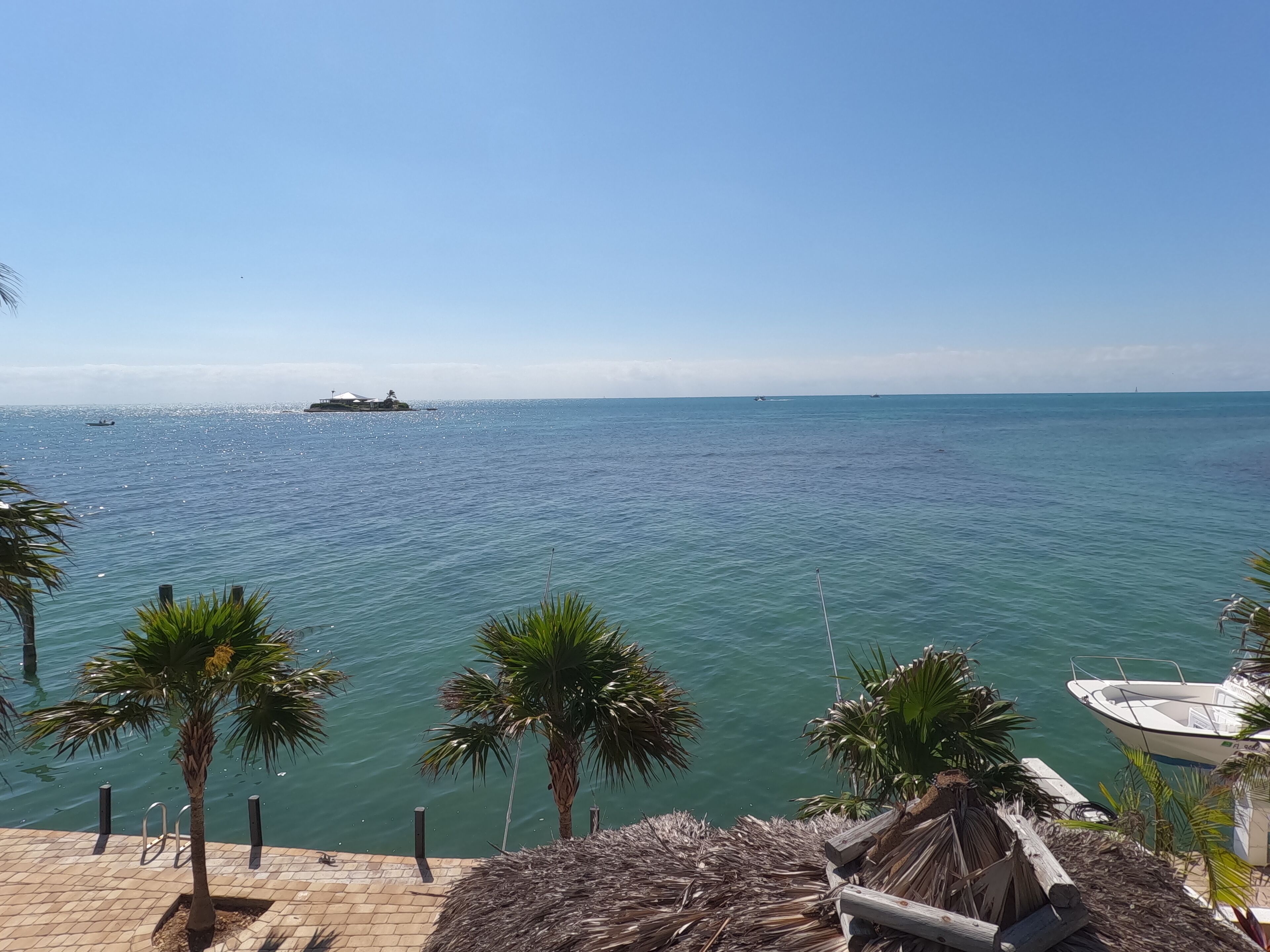 Vista de un mar tranquilo con un barco amarrado a la derecha, palmeras en primer plano y cielo despejado.
