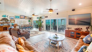 A spacious living room with a large couch, a coffee table, and a television. There are ceiling fans, a dining area, and a view of the ocean through large sliding doors. - Waterfront Oasis - 3/2 w/ Heated Pool & Direct Ocean Views (Marathon)