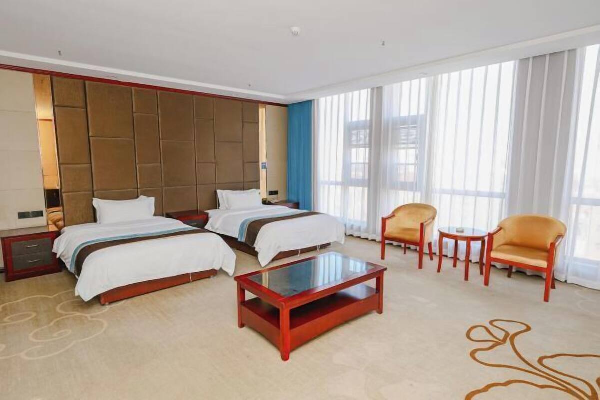 A hotel room with two beds, a glass coffee table, a small table with chairs, and large windows with curtains.