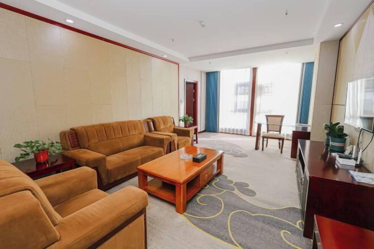 A hotel room with a sofa set, a wooden coffee table, a desk with a computer, and a large window with curtains.