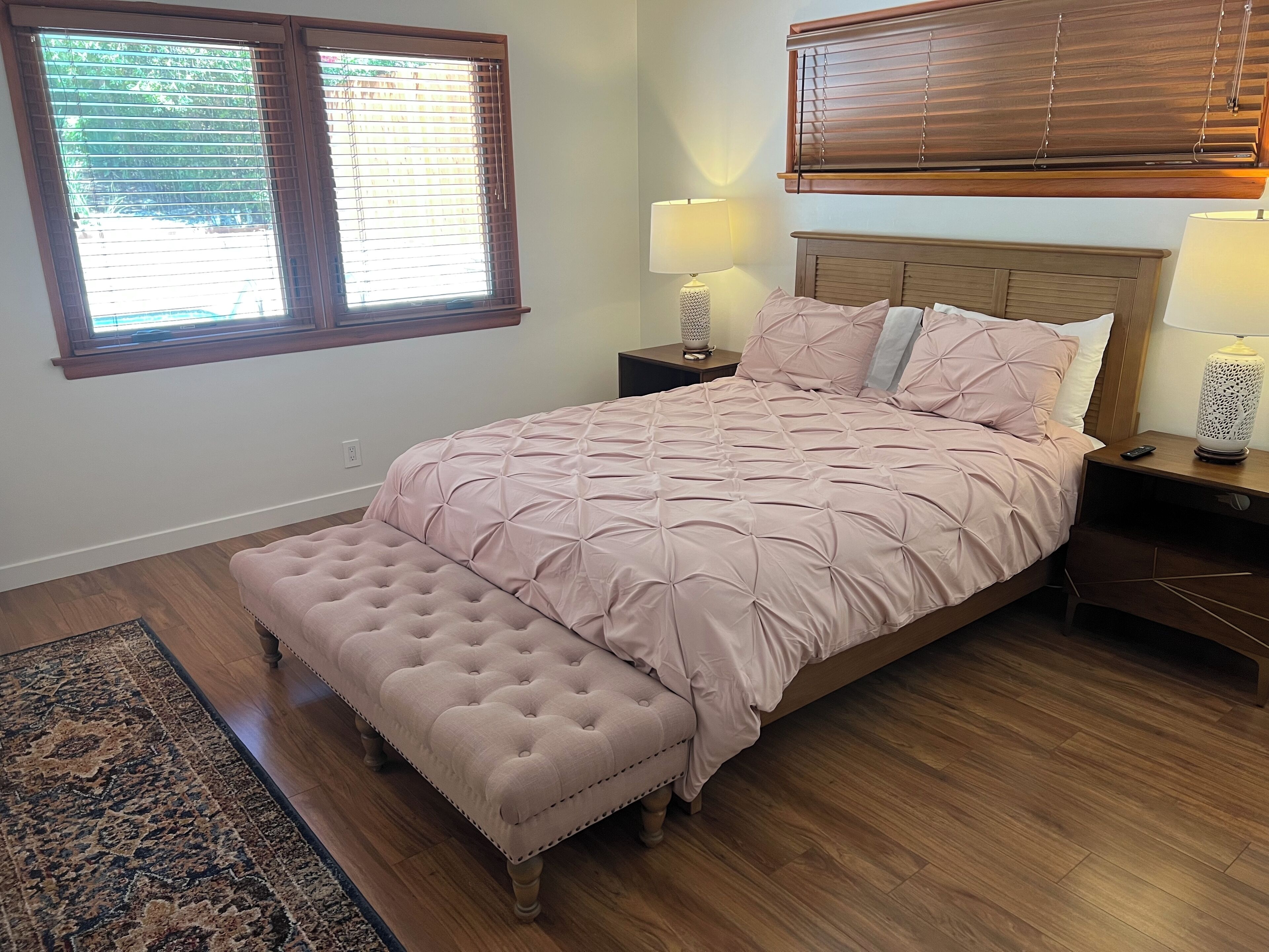 A bedroom with a large bed, a nightstand, two bedside lamps, a window with blinds, and a wooden floor.