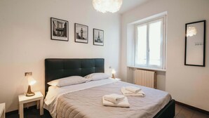 Una cama bien tendida con ropa blanca, una lámpara de noche y un cuadro con la Torre Eiffel en la pared.