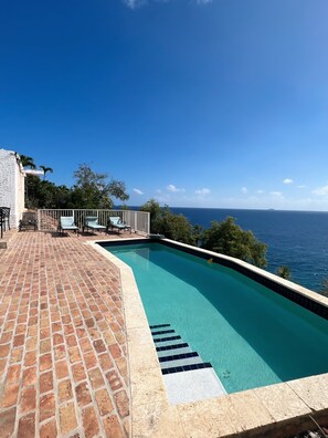 A swimming pool with a clear view of the ocean, surrounded by a brick patio and outdoor furniture. - Secluded Oceanfront Villa with pool and incredible view! Welcome to Paradise! (St John)
