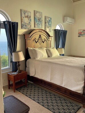 A hotel room with a bed, a nightstand, a lamp, a window with curtains, and artwork on the walls. - Secluded Oceanfront Villa with pool and incredible view! Welcome to Paradise! (St John)