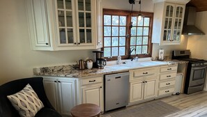 Une cuisine avec des meubles blancs, un plan de travail en granit, un lave-vaisselle, une cuisinière et une fenêtre donnant sur les arbres.