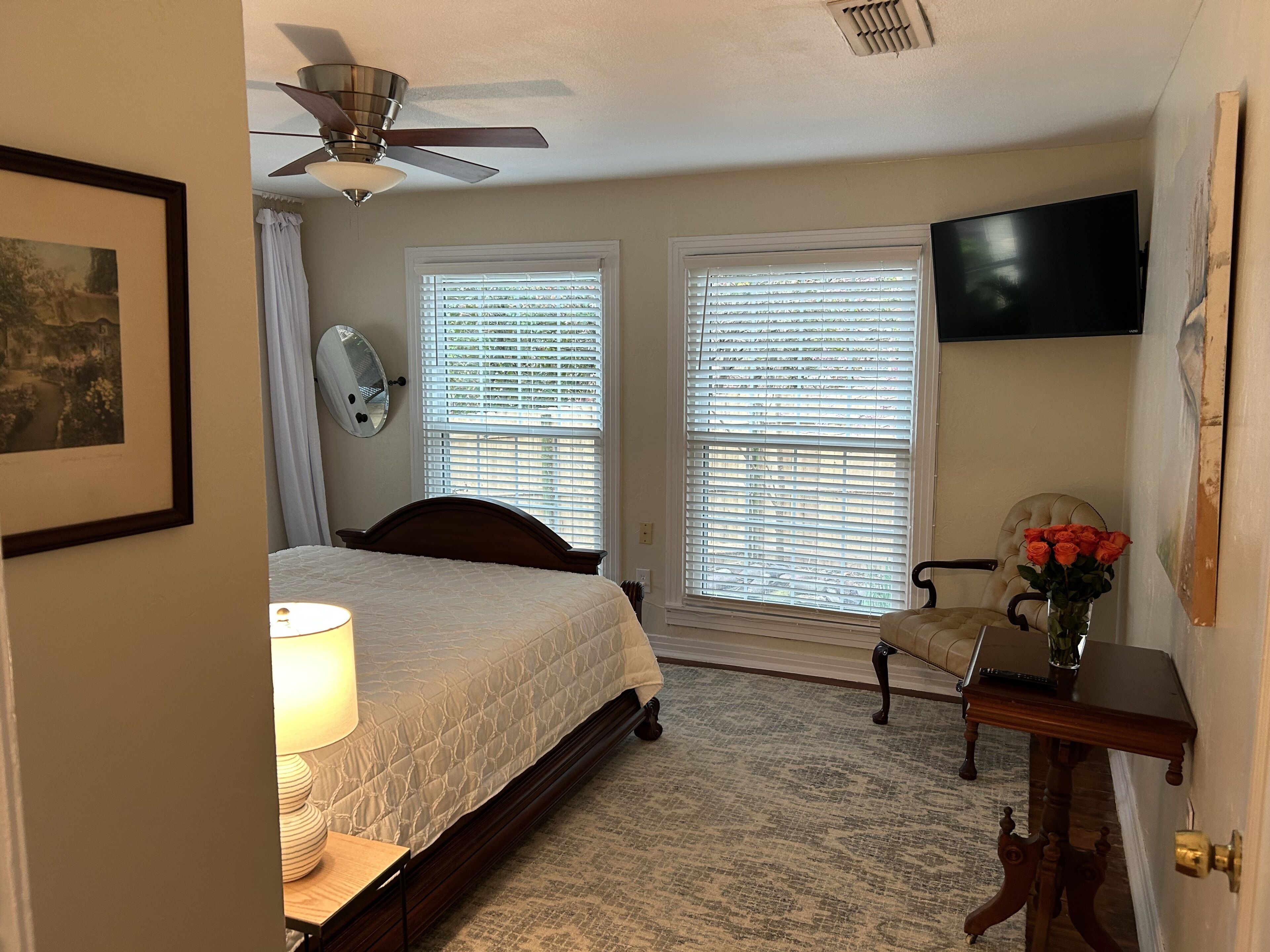 A bedroom with a bed, a nightstand, a lamp, a chair, a desk, a vase of flowers, a ceiling fan, and a TV mounted on the wall.