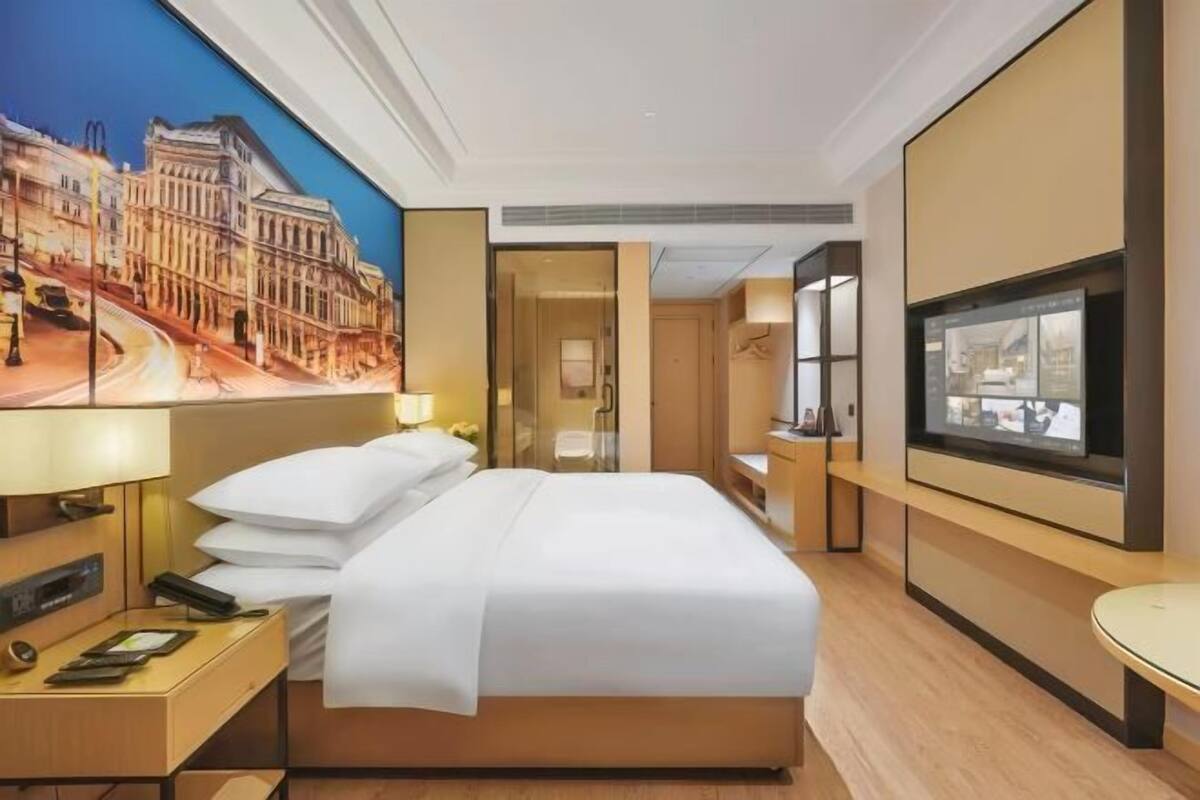 A hotel room with a large bed, a desk with a phone and a tablet, a TV, and a painting of a cityscape.