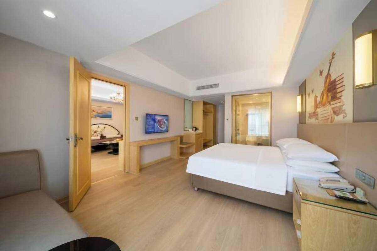 A hotel room with a bed, bedside table, and a desk with a chair. There is a door leading to another room.