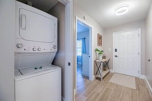 Uma lavanderia com máquina de lavar e secadora, um corredor com porta para outro cômodo e uma pequena mesa com uma planta.