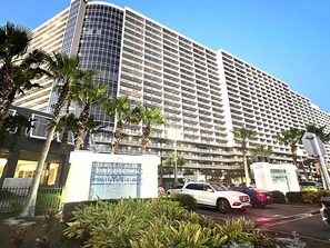 Hotel com várias andares, fachada curva em vidro, entrada com portão e área ajardinada com palmeiras.