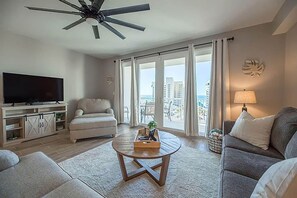 Una sala de estar con televisor de pantalla plana, ventilador de techo, sofá, una mesa de centro y vista a la ciudad a través de grandes ventanales.