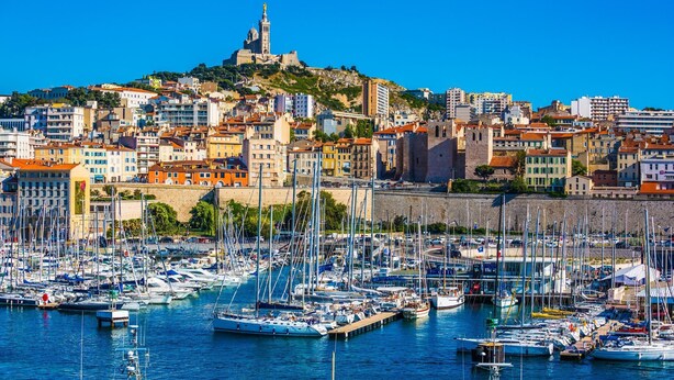 Puerto deportivo lleno de barcos, una colina histórica con una torre y un paisaje urbano con edificios.