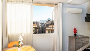 Habitación de hotel con vistas a un paisaje urbano, una mesa de comedor con una orquídea en maceta, un mueble bajo y un equipo de aire acondicionado.