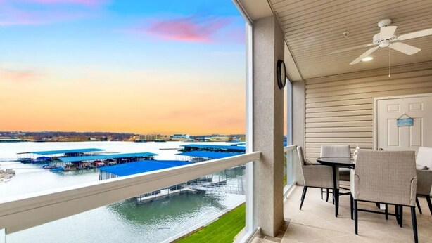 Uma varanda com vista para um porto e barcos, uma área de refeições com cadeiras e mesa, e um ventilador de teto.