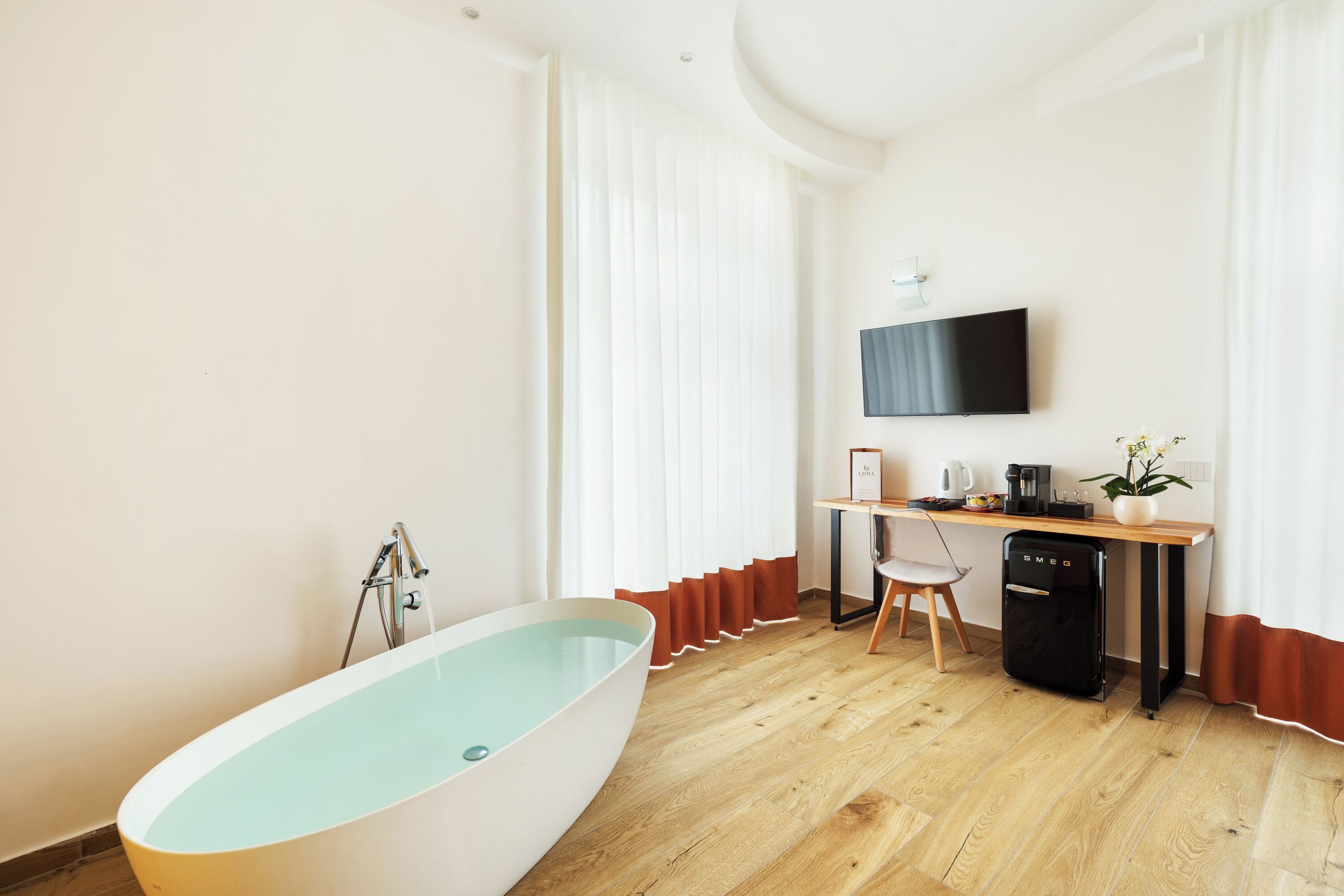 A modern bathroom with a freestanding bathtub, a wooden desk, a black dishwasher, and a mounted TV.