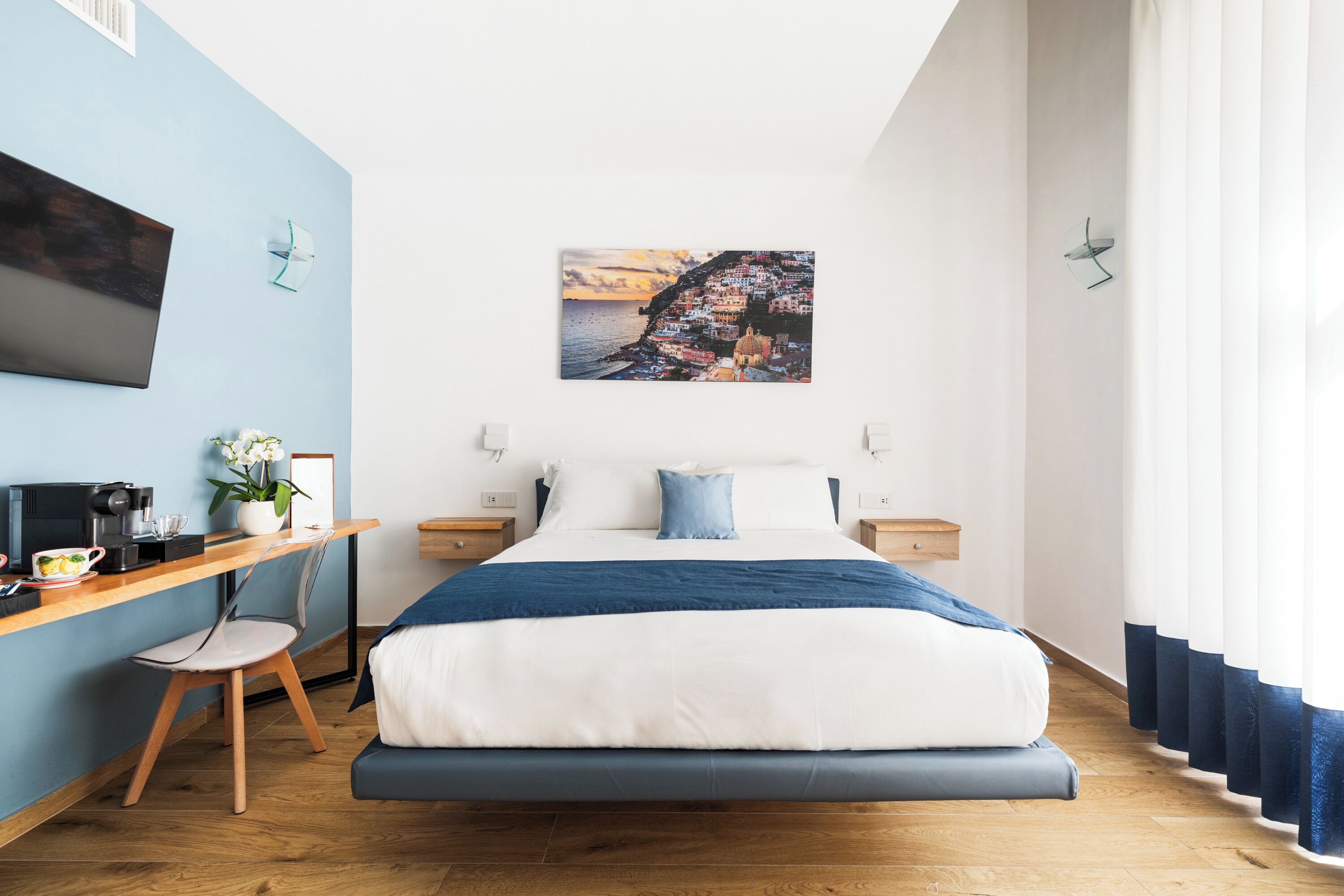 A modern hotel room with a bed, a desk with a coffee maker, a chair, and a painting of a coastal town on the wall.