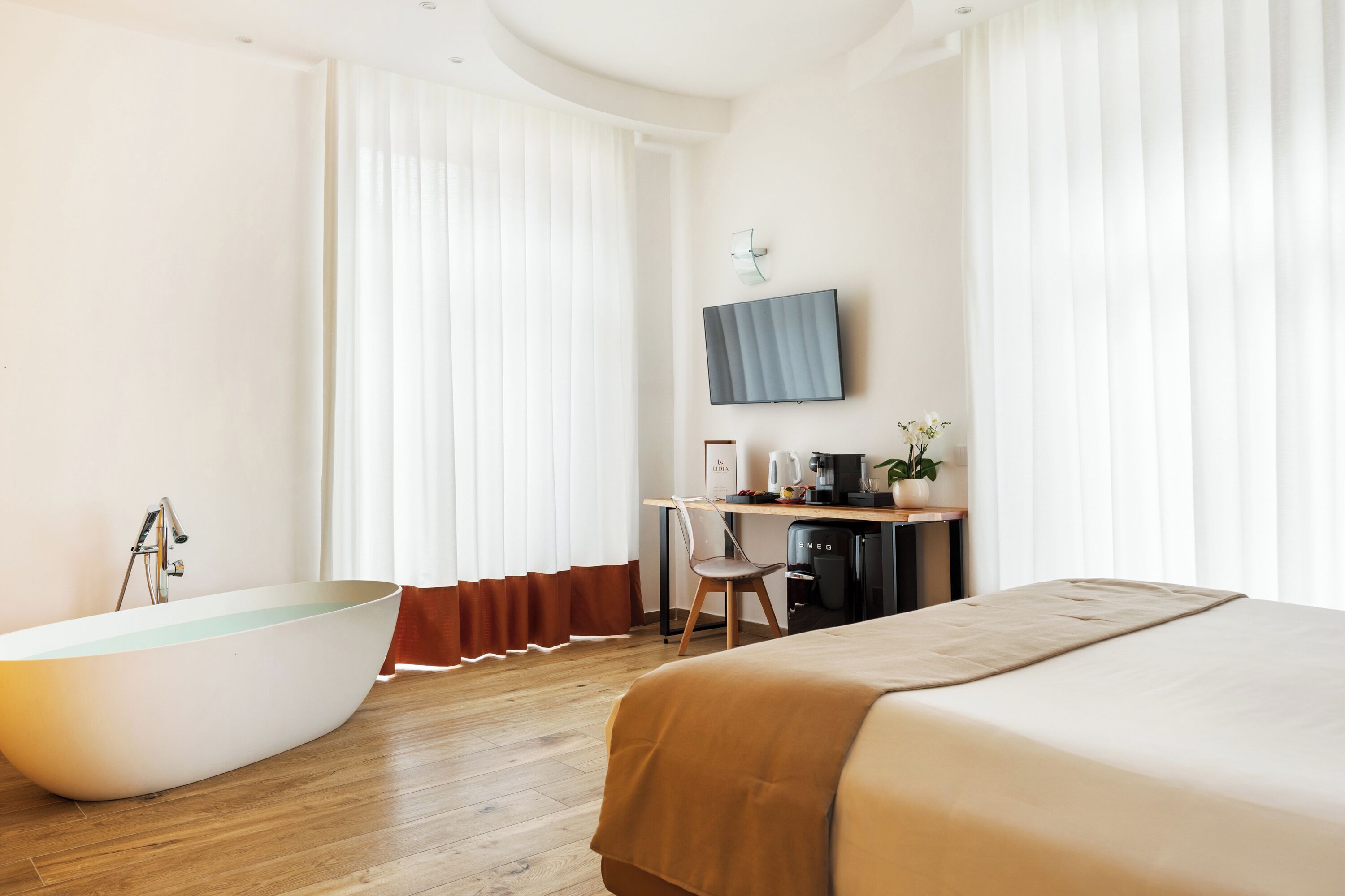 Une chambre d’hôtel moderne dotée d’une grande baignoire, d’un lit, d’un bureau avec un ordinateur et d’un téléviseur fixé au mur.