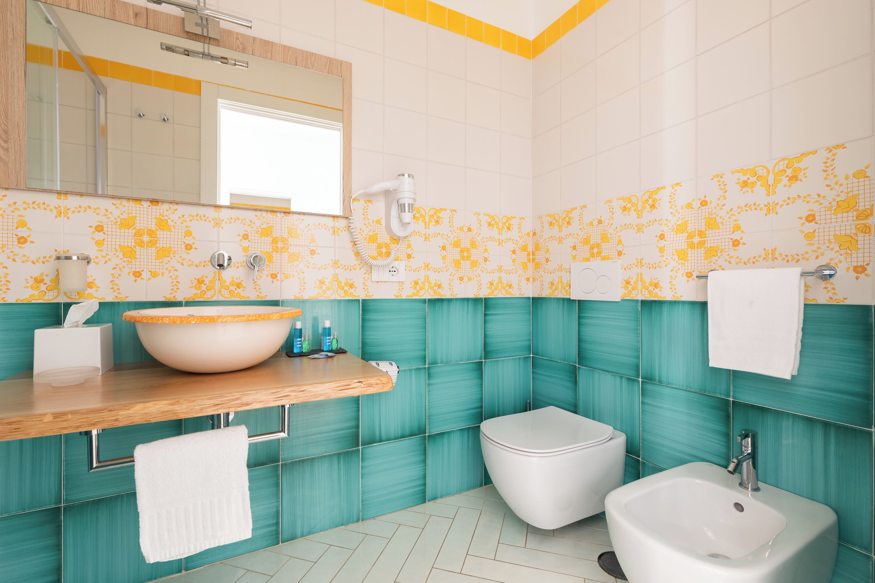 Ein Badezimmer mit großem Waschbecken, Toilette und Bidet, ausgestattet mit Spiegel, Handtuchhalter und gefliesten Wänden.