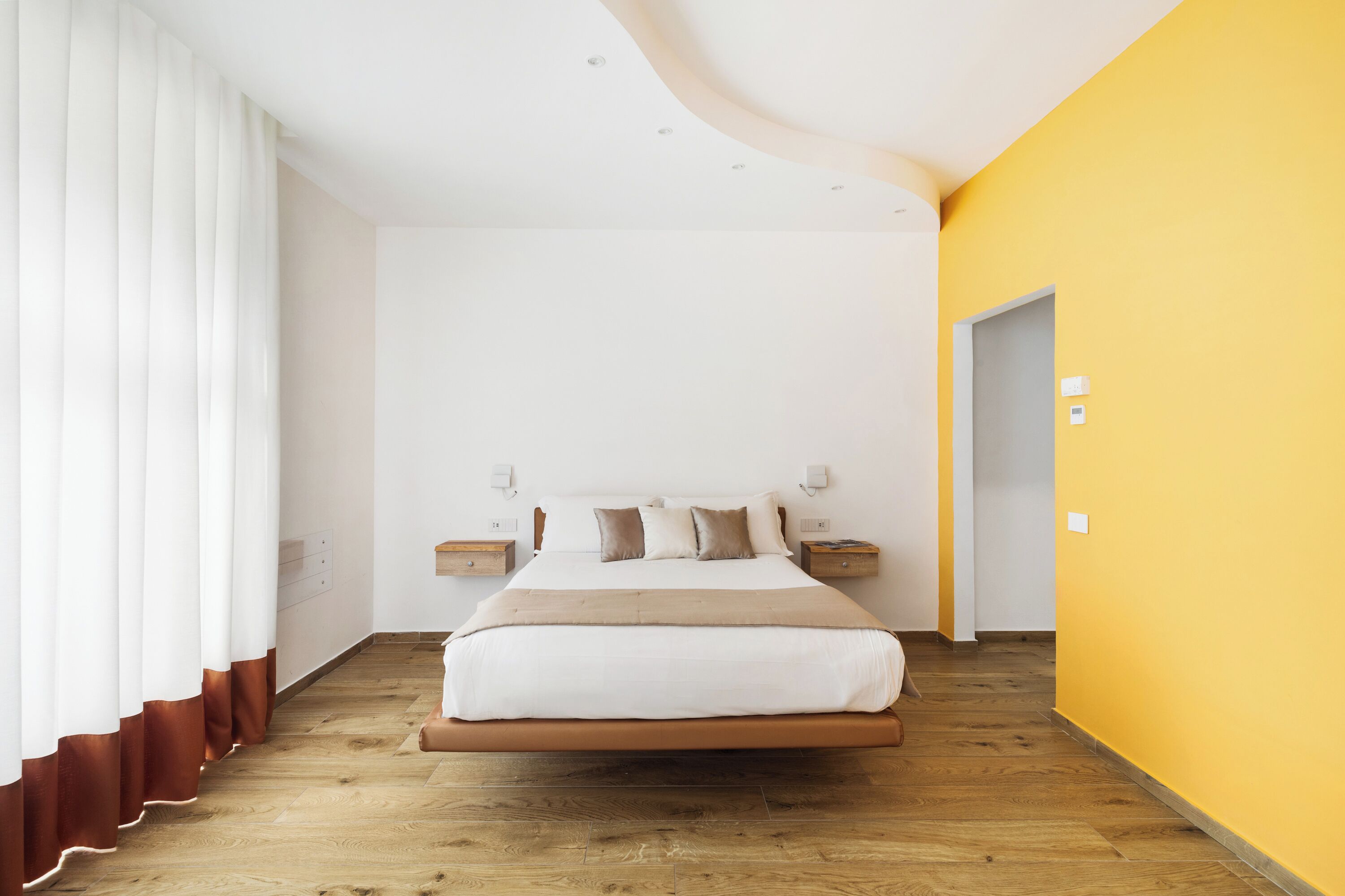 Une chambre moderne avec un grand lit, un plancher en bois, des murs blancs et un mur d’accent jaune.