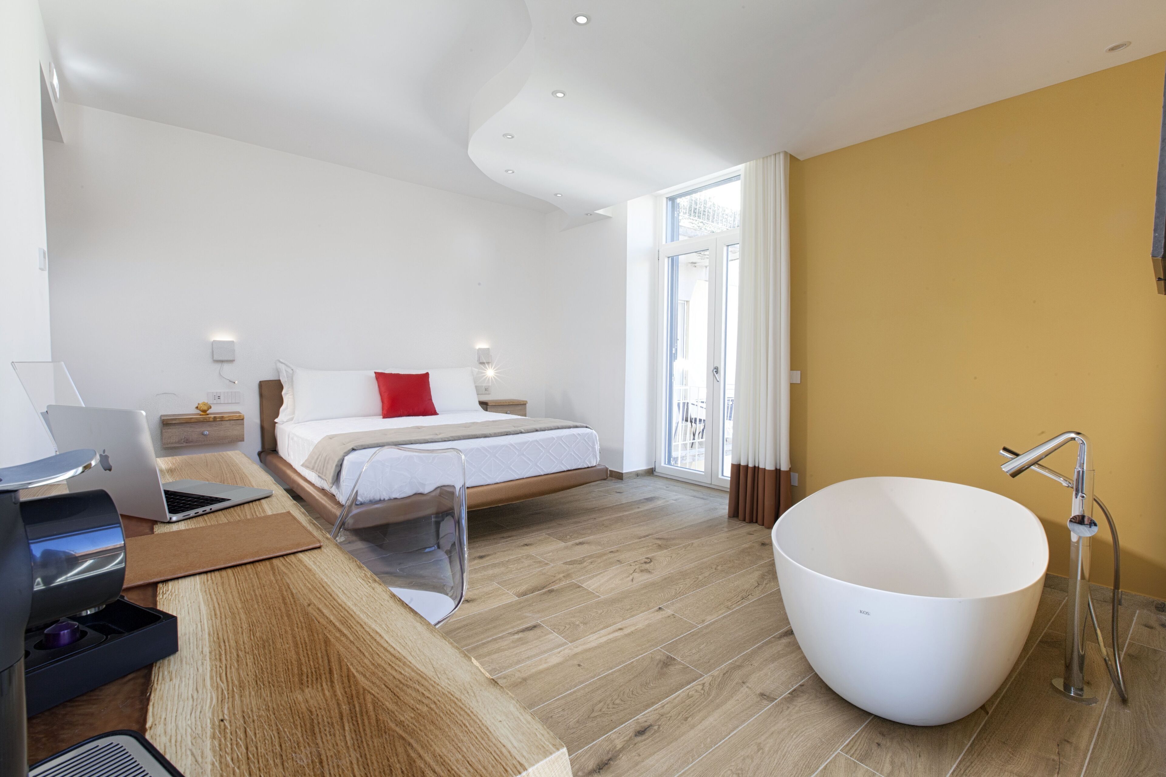 Ein modernes Hotelzimmer mit einer großen weißen Badewanne, einem Holztisch mit einem Laptop, einem Bett mit weißer Bettwäsche und einem roten Kopfkissen sowie einem großen Fenster mit Vorhängen.