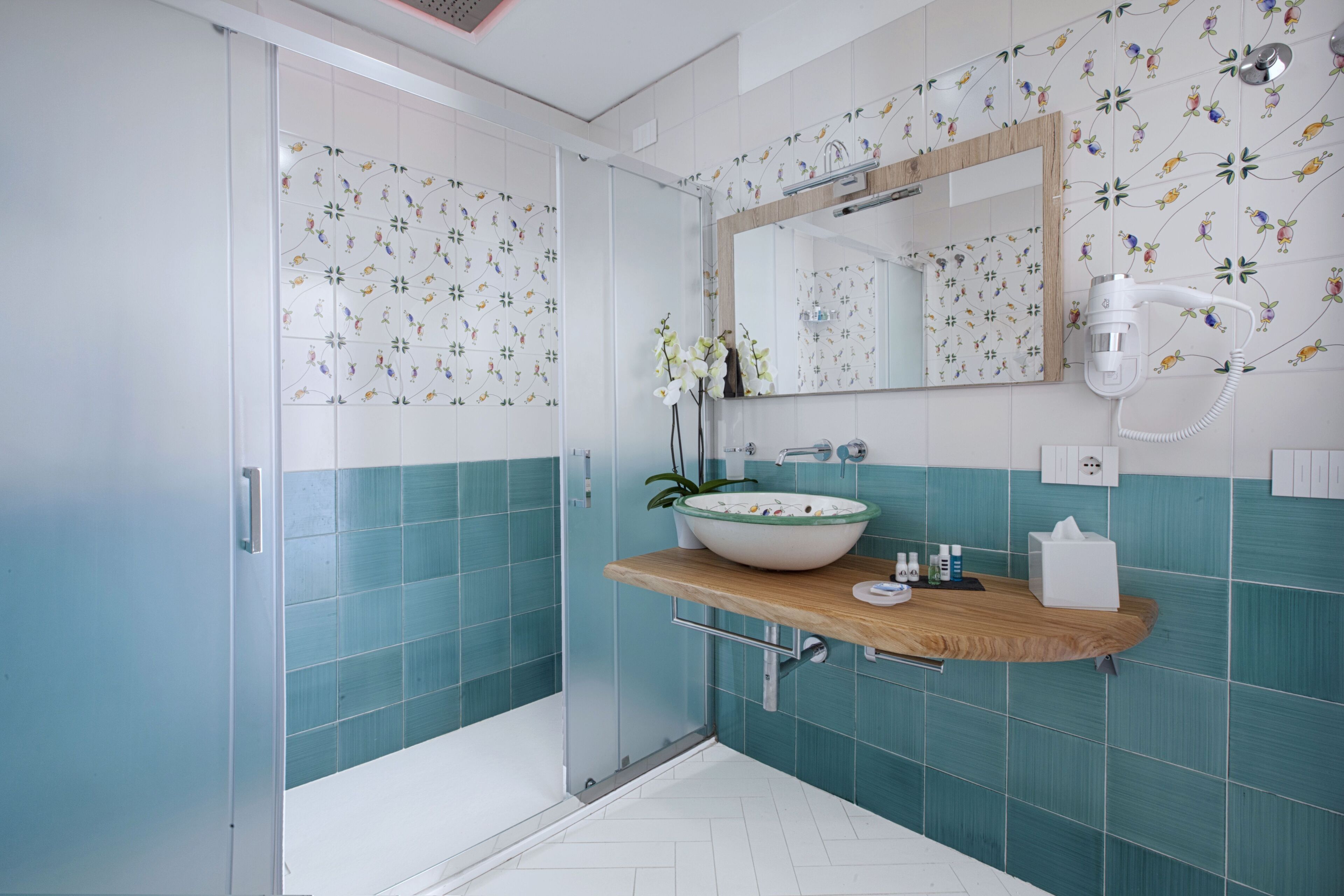 A bathroom with a glass shower enclosure, a wooden vanity with a bowl sink, and a mirror with a floral tile backsplash.
