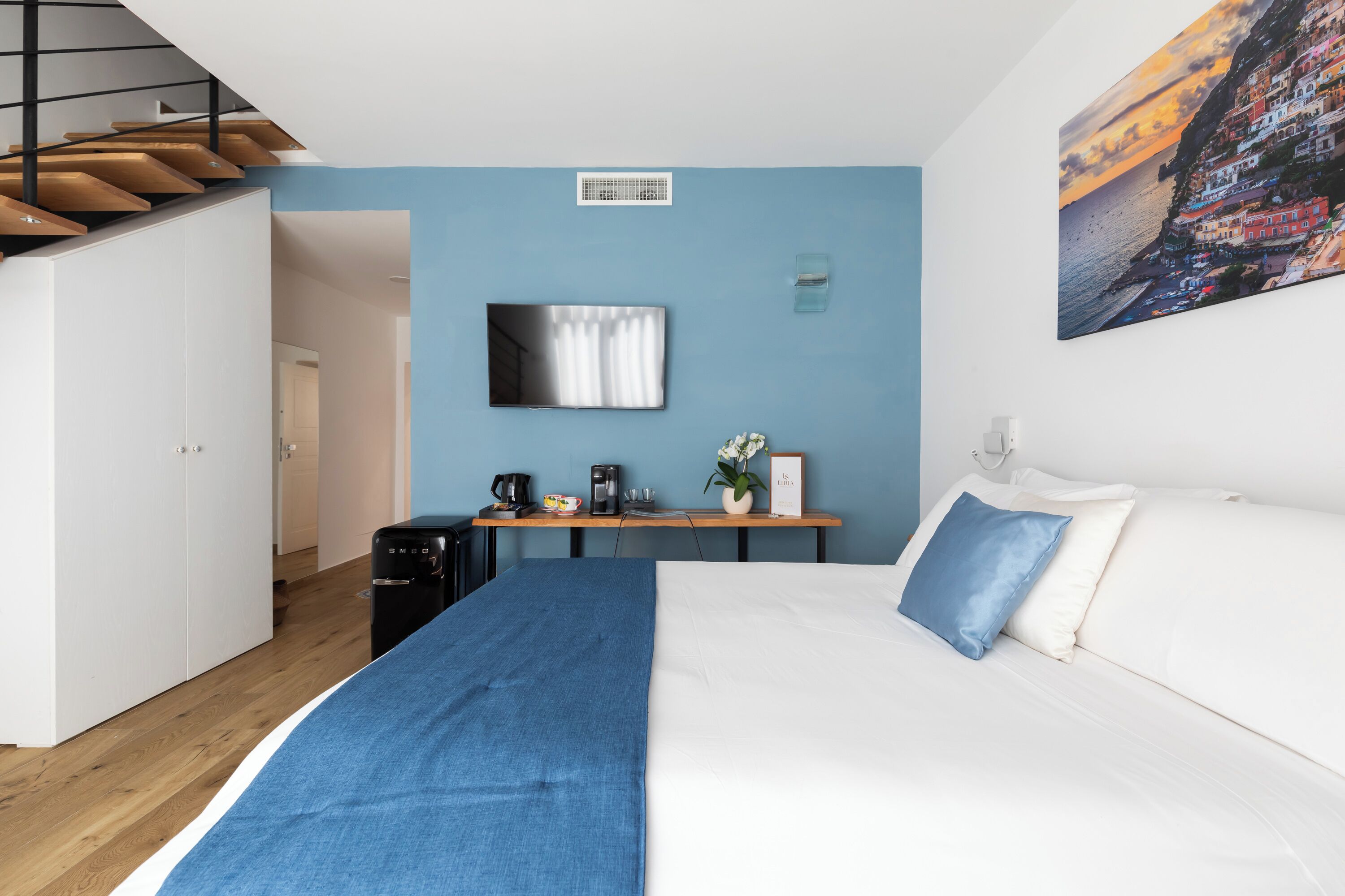 A bedroom with a bed, a desk with a coffee machine, a TV, and a painting of a coastal scene.