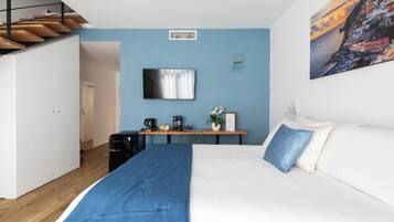 A bedroom with a bed, a desk with a coffee machine, a TV, and a painting of a coastal scene.