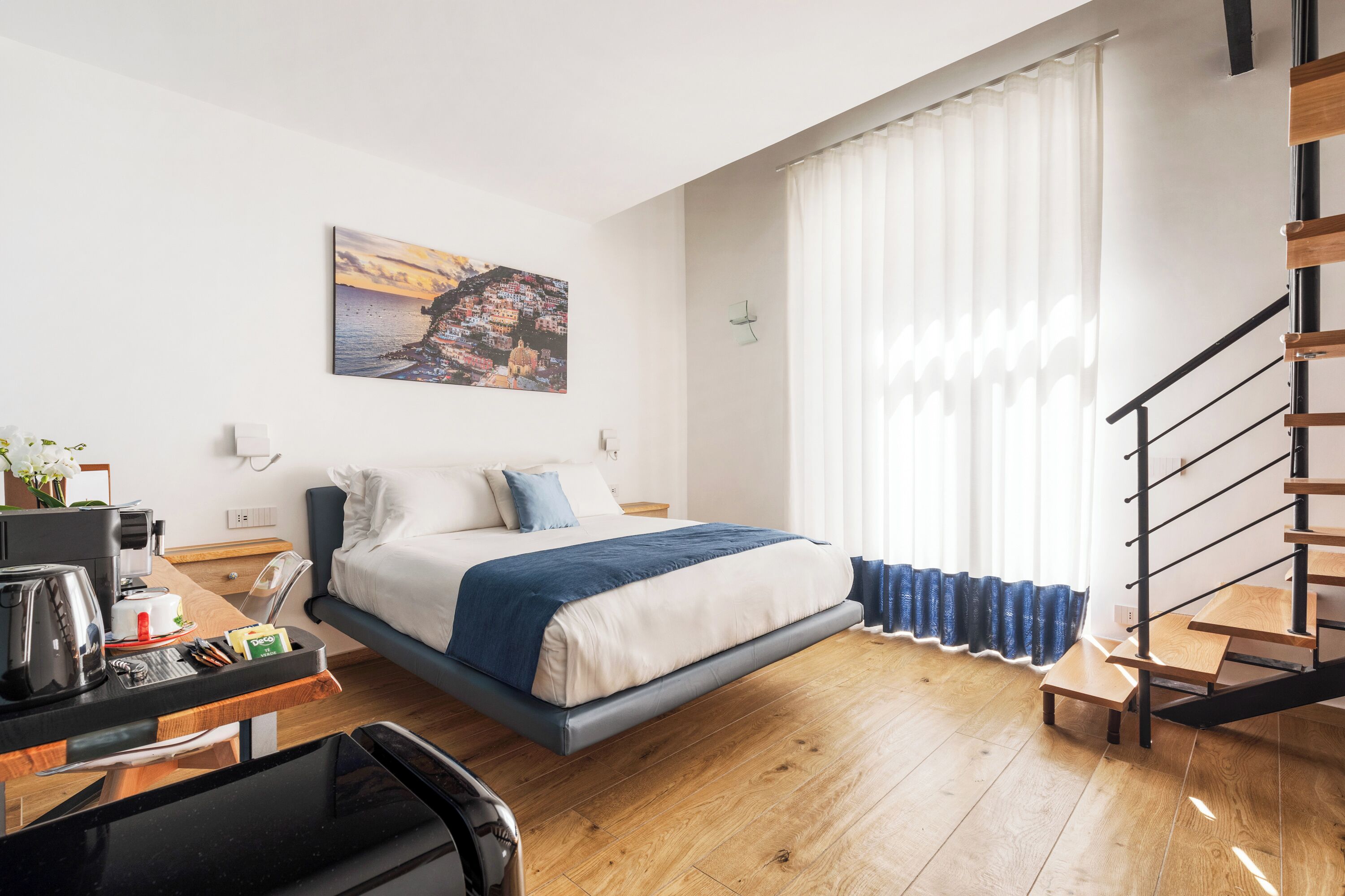 Une chambre d’hôtel moderne équipée d’un lit, d’un bureau avec une machine à café, d’une chaise et offrant une vue sur une ville côtière par la fenêtre.
