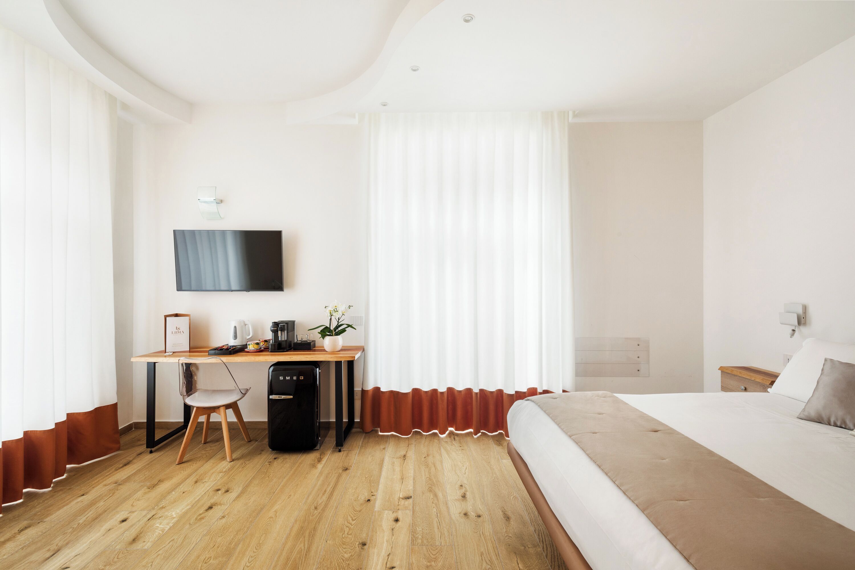 A hotel room with a wooden desk, a bed, a TV mounted on the wall, and a window with curtains.