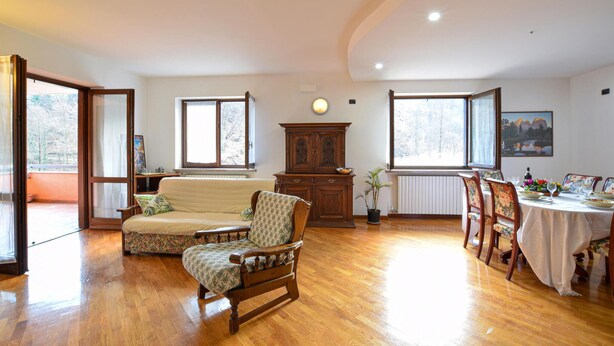 A spacious living room with a wooden floor, a sofa, a wooden chair, a dining table set for four, and a large window with a view of trees.