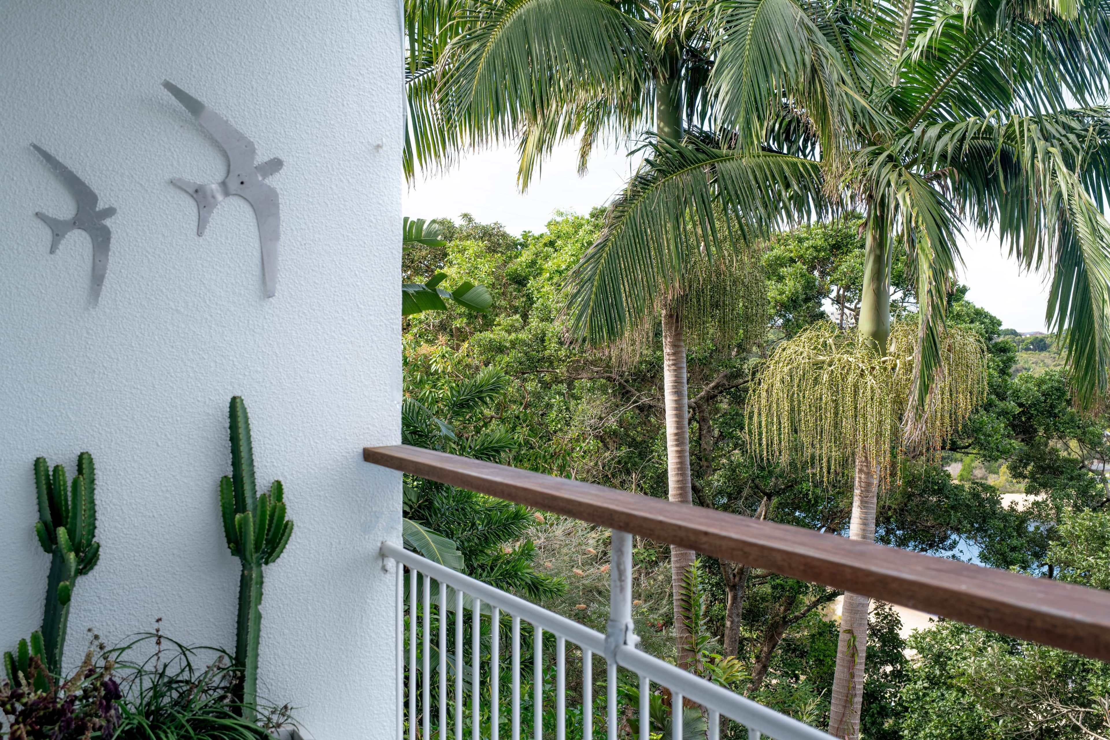 Een balkon met een witte reling, cactussen en uitzicht op een weelderig groen landschap met palmbomen.