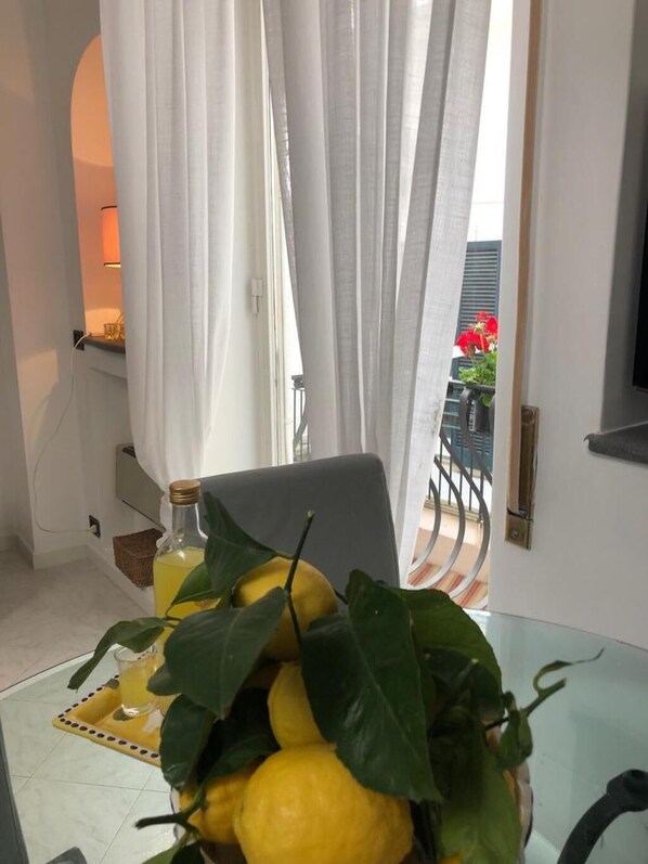 Une pièce avec une table en verre sur laquelle sont posés des citrons et une bouteille, des rideaux blancs et une vue sur un balcon orné de fleurs rouges.