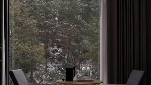 Une pièce avec une grande fenêtre, deux chaises, une petite table ronde sur laquelle se trouve un objet noir et un récipient en verre.
