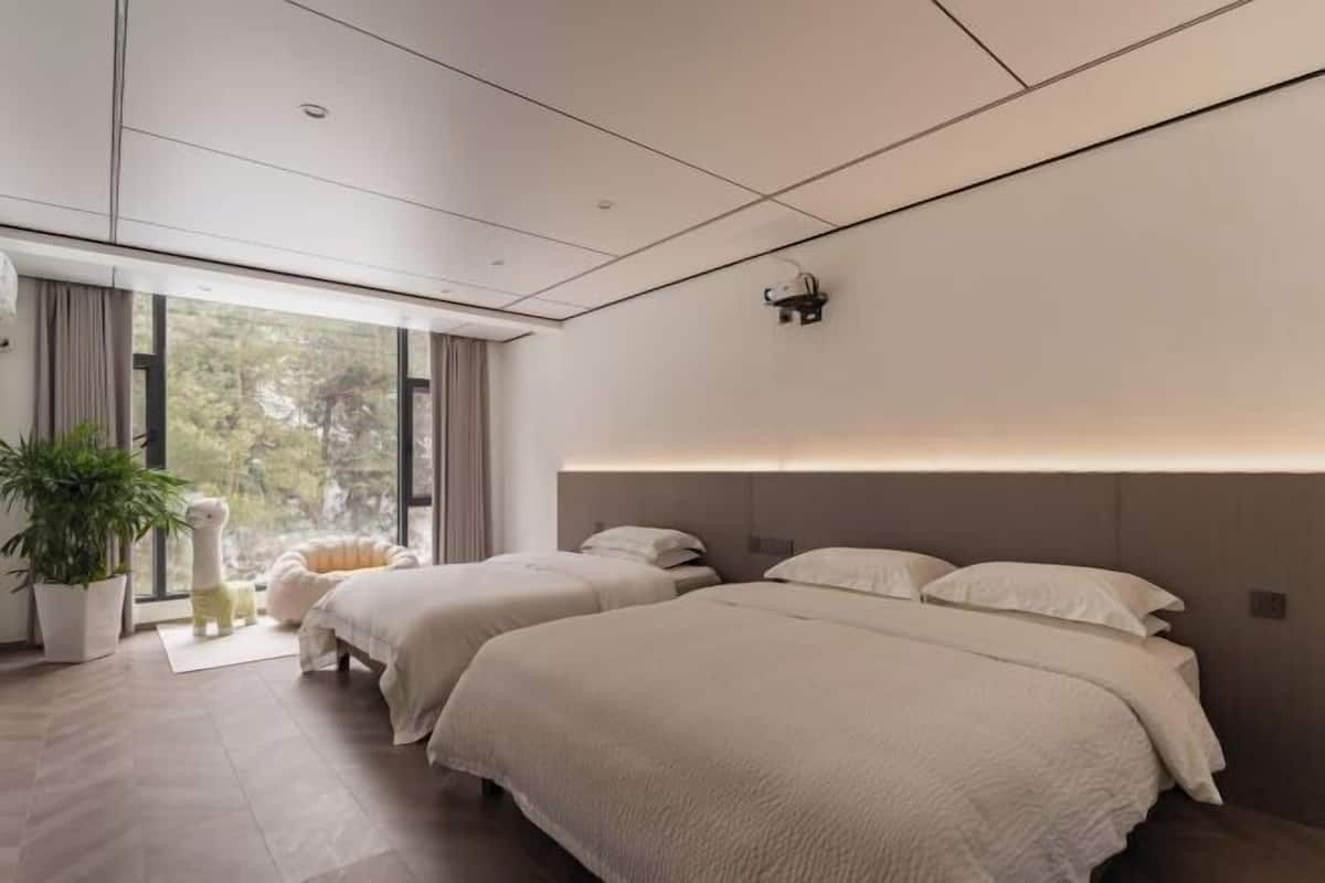 Een moderne hotelkamer met twee bedden, een groot raam, een plant in pot en een comfortabele fauteuil.
