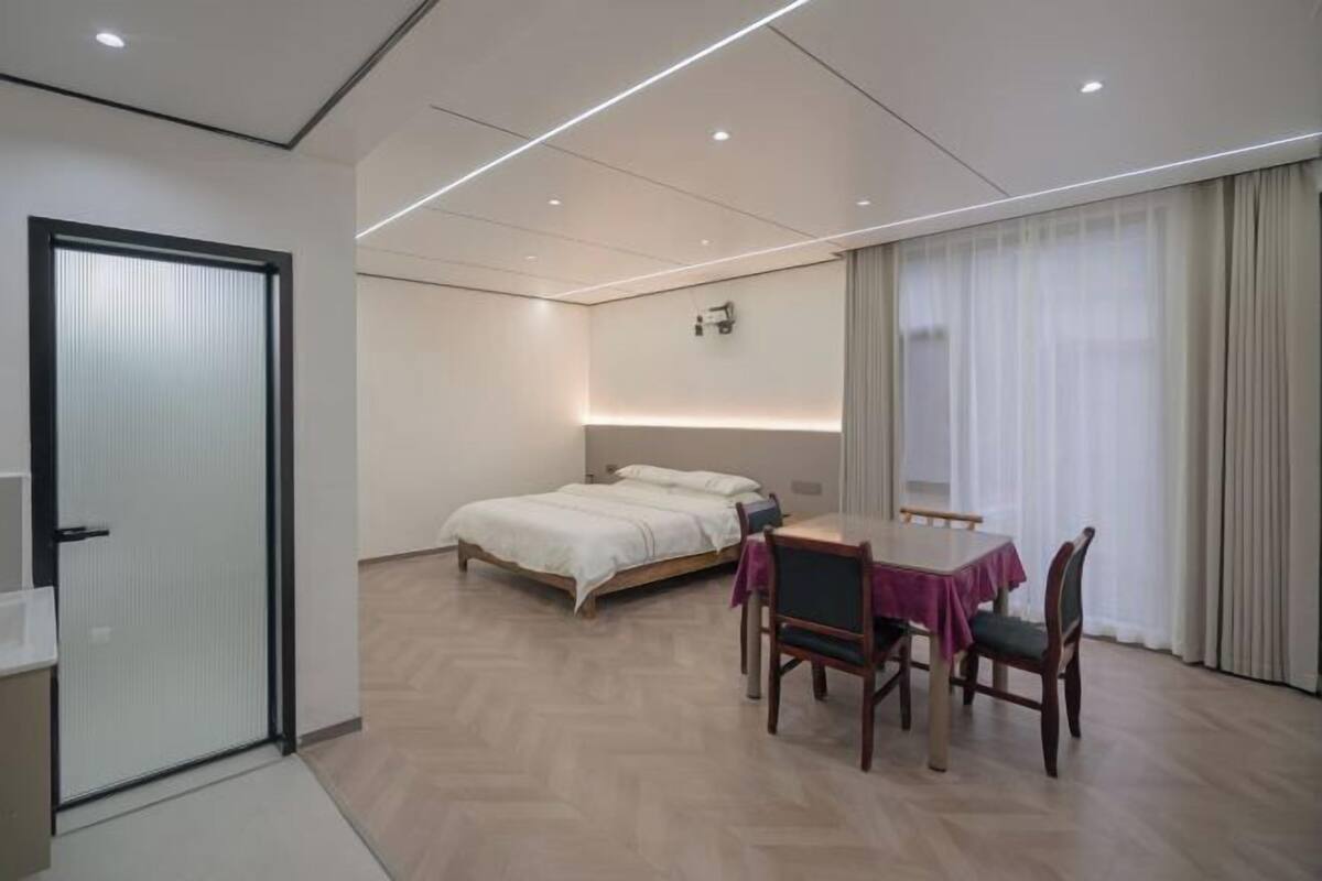Een moderne hotelkamer met een bed, een eettafel met stoelen, een klein bureau, een raam met gordijnen en een deur.
