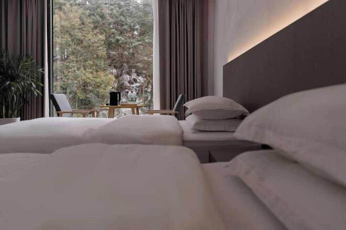 Een slaapkamer met een bed, een nachtkastje, een stoel en uitzicht op bomen door grote ramen.