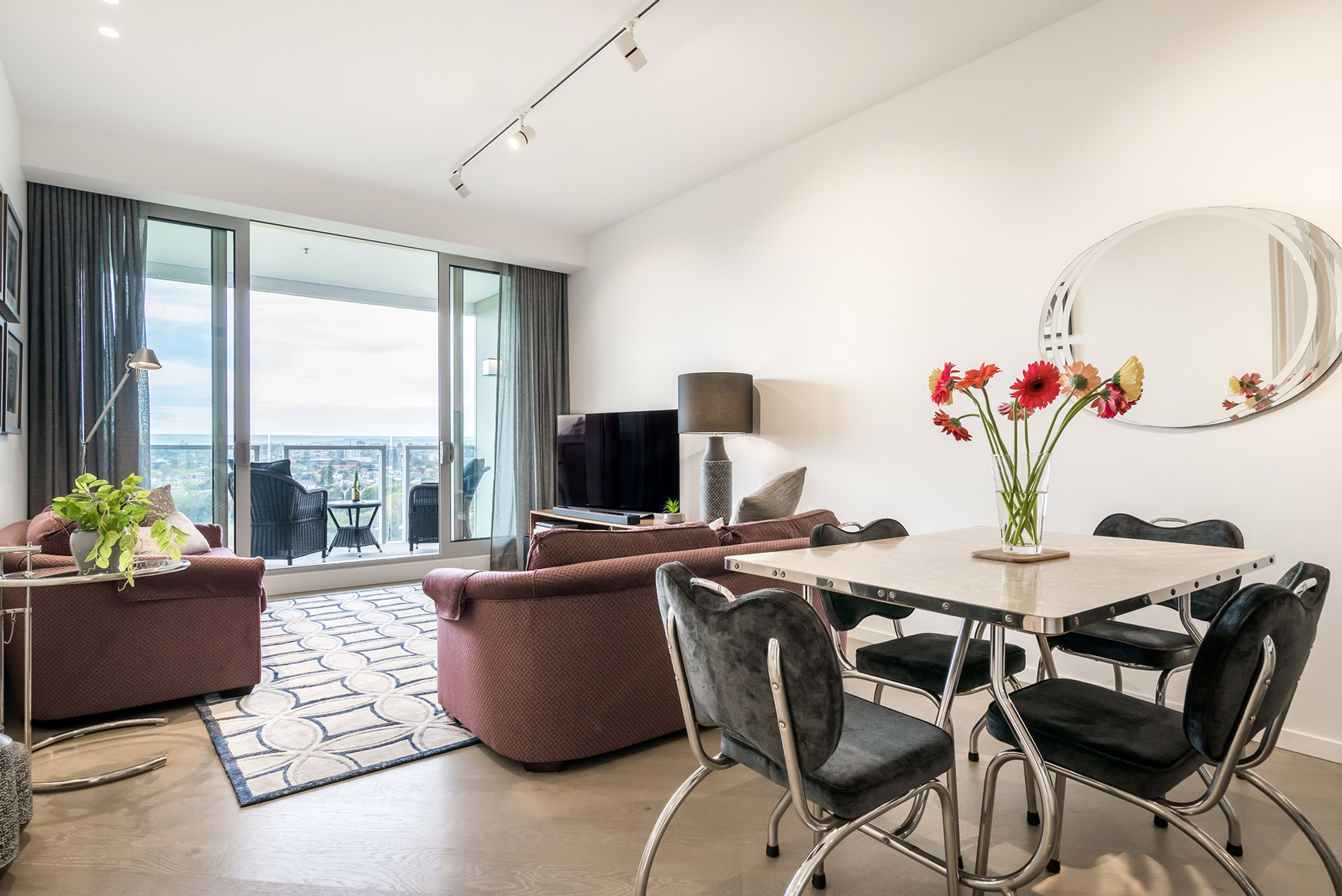 A modern living room with a sofa, a dining table with chairs, a flat-screen TV, and a large window offering a city view.