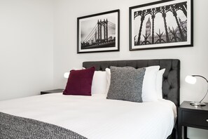 A neatly made bed with a white duvet, two pillows, and a grey throw blanket. There are two framed pictures on the wall, one of a bridge and the other of a tree silhouette. - The Fawkner Stylish Saint Kilda Apartment (Melbourne)