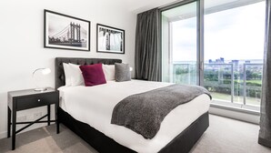 Une chambre moderne avec un grand lit, une table de chevet, deux tableaux encadrés, une fenêtre donnant sur la ville et un rideau gris.