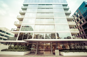 A modern multi-story building with a glass facade, a central entrance, and a landscaped area with plants. - The Fawkner Stylish Saint Kilda Apartment (Melbourne)